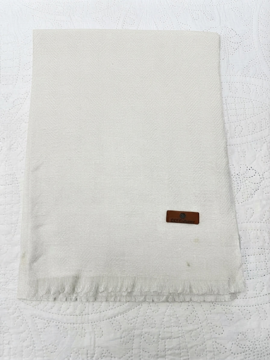 An off-white woven fabric napkin with fringed edges and a small brown leather brand tag, laid flat on a white textured lace tablecloth.