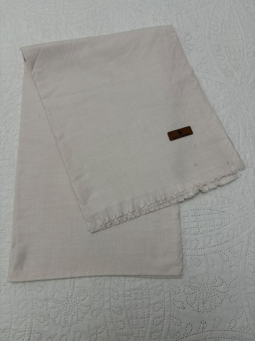 A folded light beige linen napkin with a small brown leather brand tag, displayed on a white textured quilted tablecloth.