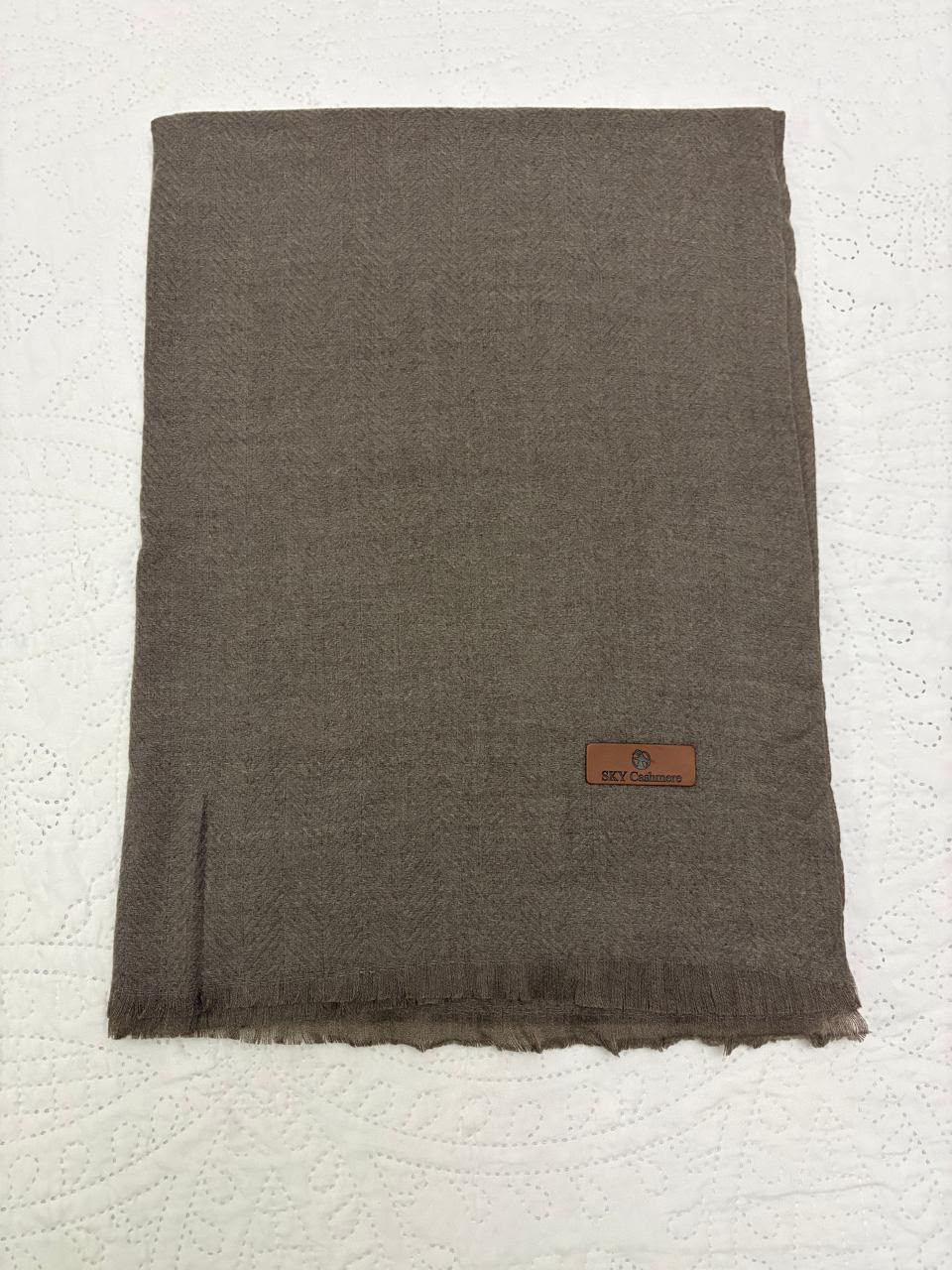 A folded dark brown woven scarf with fringed edges and a small brown leather brand patch, laid flat on a white textured background.