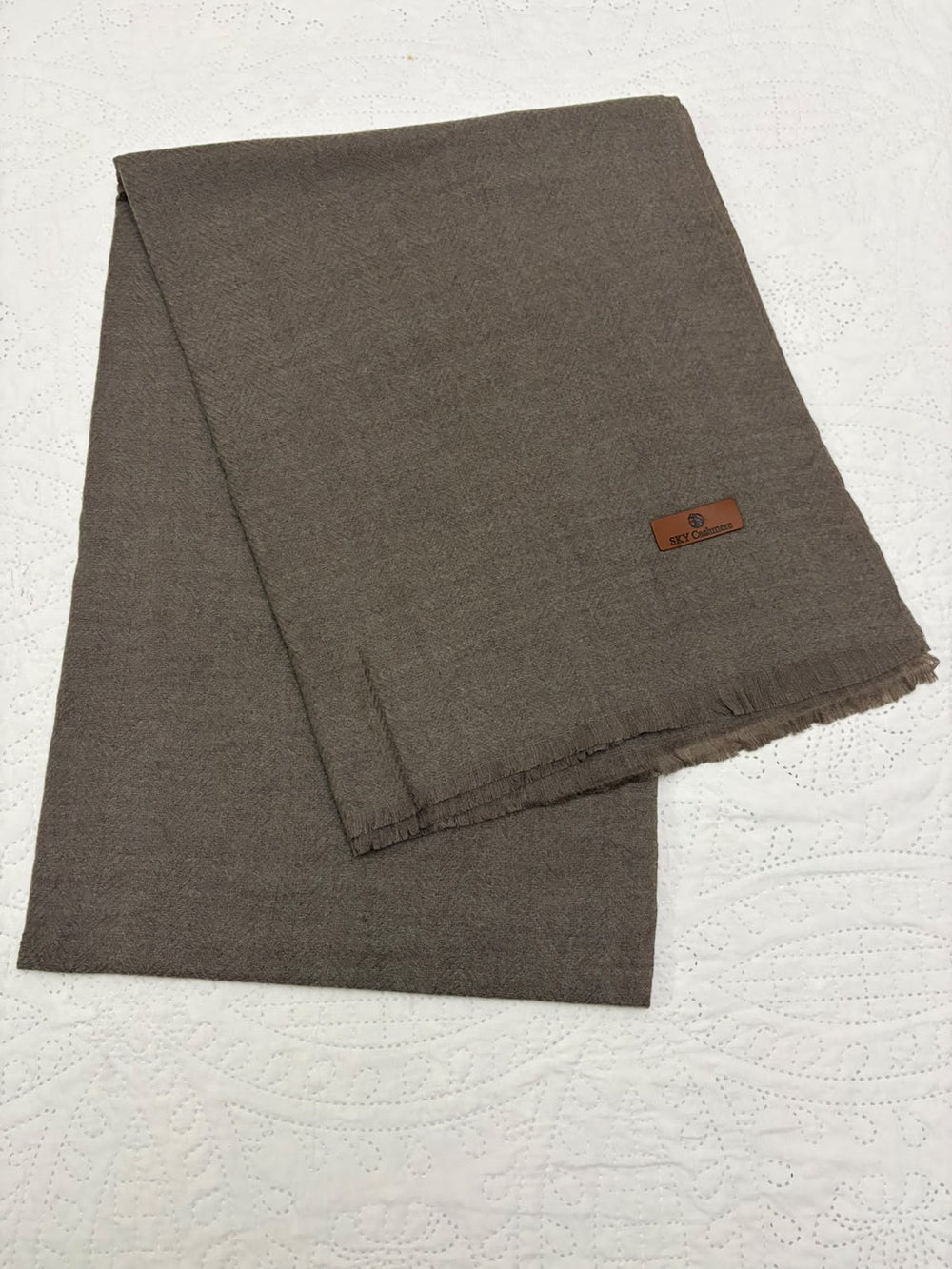 A folded dark brown woven scarf with a small brown leather brand tag, displayed on a textured white fabric background.