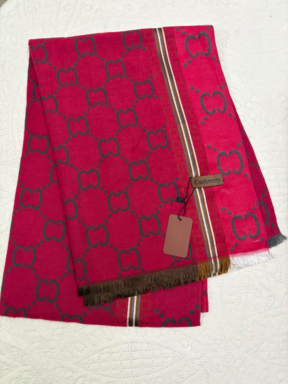 A folded Gucci scarf in vibrant pink with a dark green GG monogram pattern, featuring a striped border and a hanging brand tag on a textured white background.
