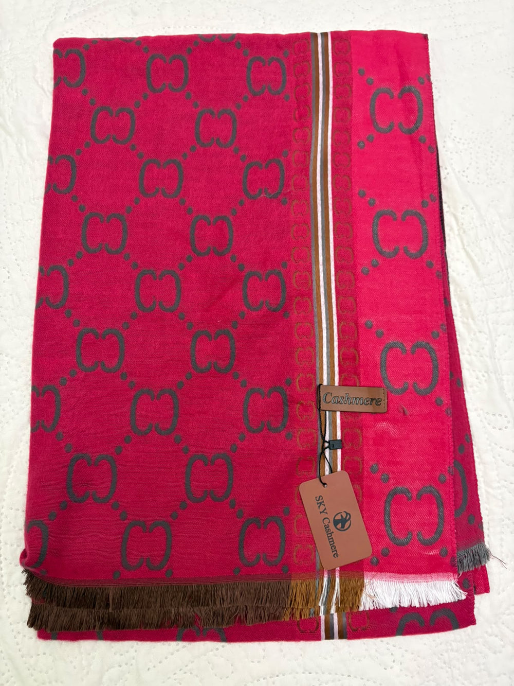A folded Gucci scarf in vibrant pink with the signature dark green GG monogram pattern, featuring a striped border and a hanging brand tag.