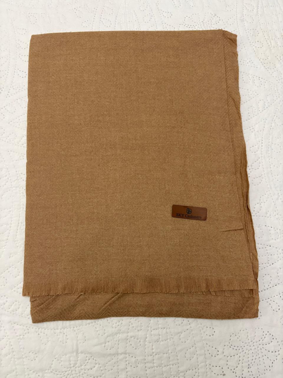 A folded brown woven scarf with fringed edges and a small dark brown brand label, laid flat on a white textured fabric background.
