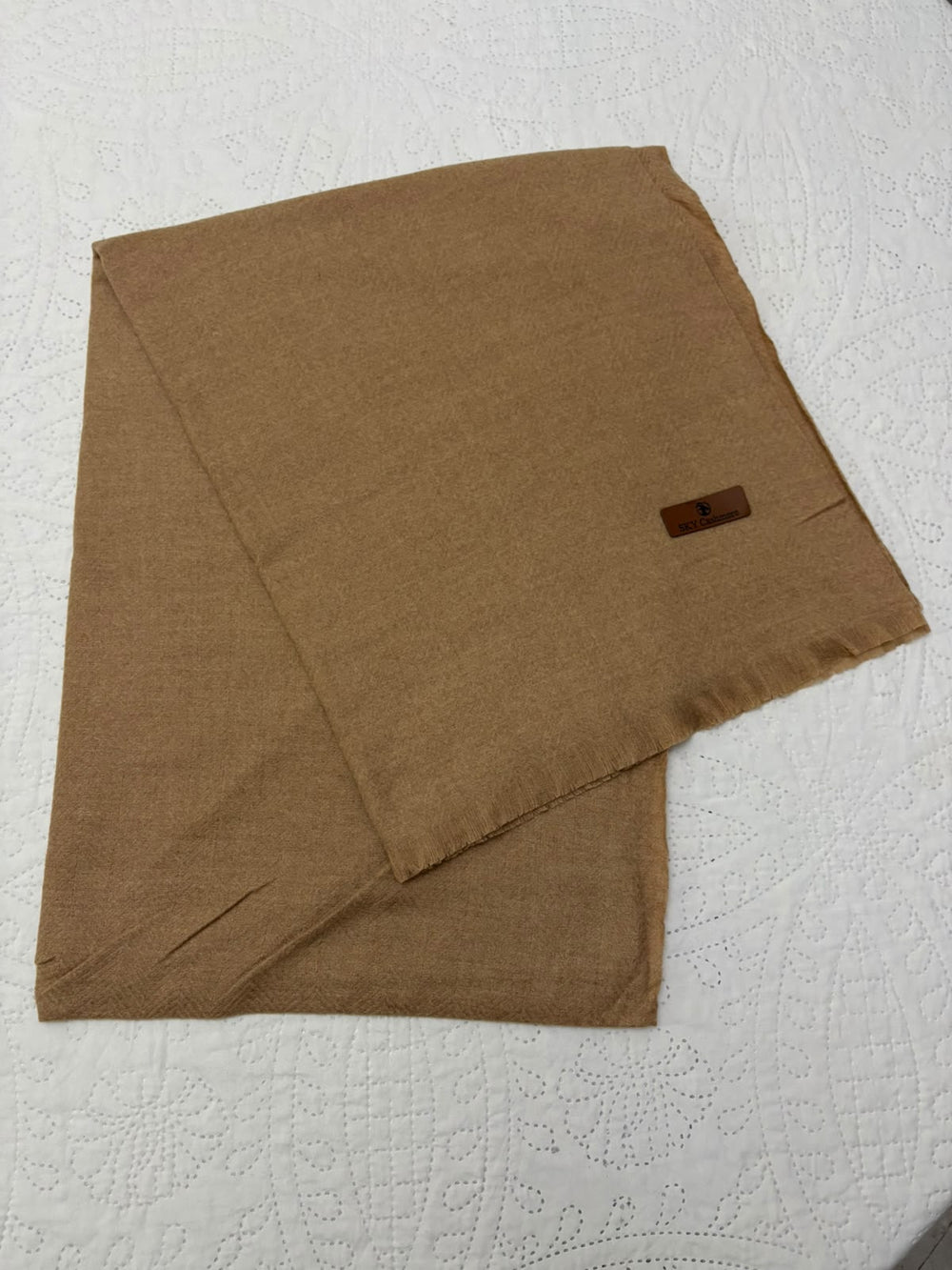 A folded brown woven scarf or shawl with a small dark brown rectangular label, laid flat on a white textured fabric background.