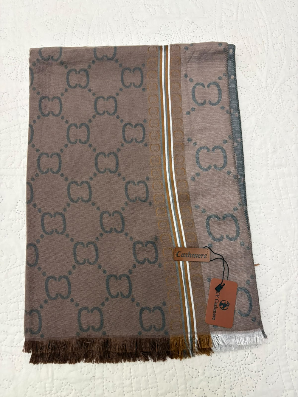 A folded Gucci scarf in taupe brown with the signature GG monogram pattern in dark gray, featuring a striped border and a brown leather brand tag with a hanging tag, laid flat on a white textured fabric background.