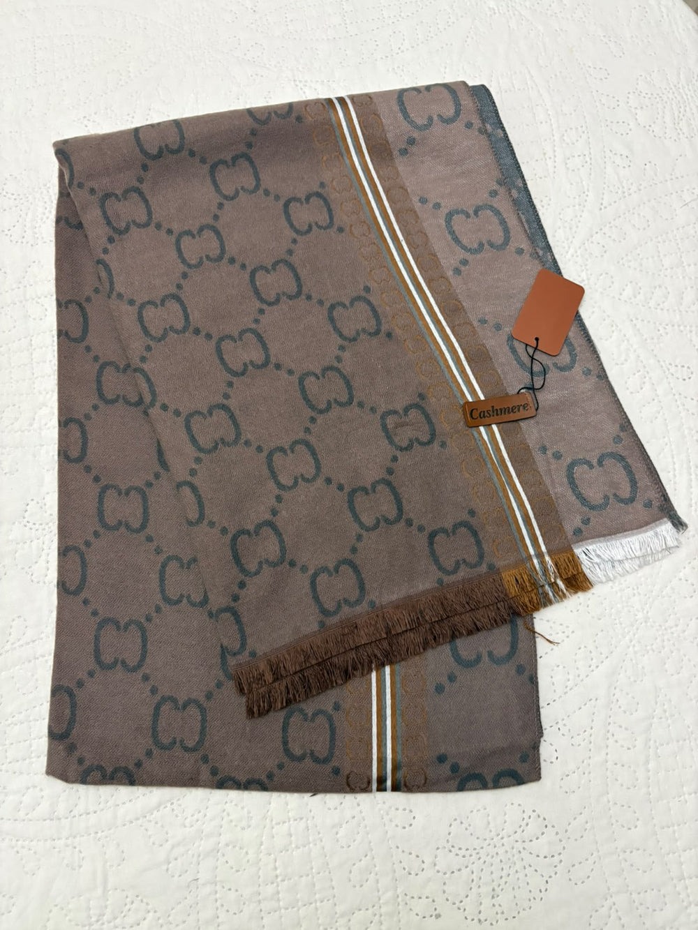 A folded Gucci scarf in taupe brown with the brand's interlocking GG monogram pattern in dark brown, featuring a striped border with white and gold accents, fringed edges, and a brown leather brand tag with a paper tag attached.