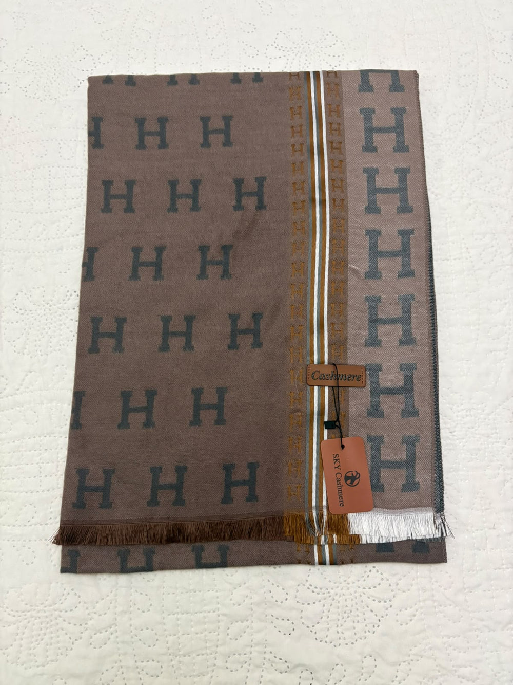 A folded Hermès scarf in taupe brown with a repeating dark gray 'H' pattern, featuring a central gold and white stripe border and a hanging orange Hermès tag.
