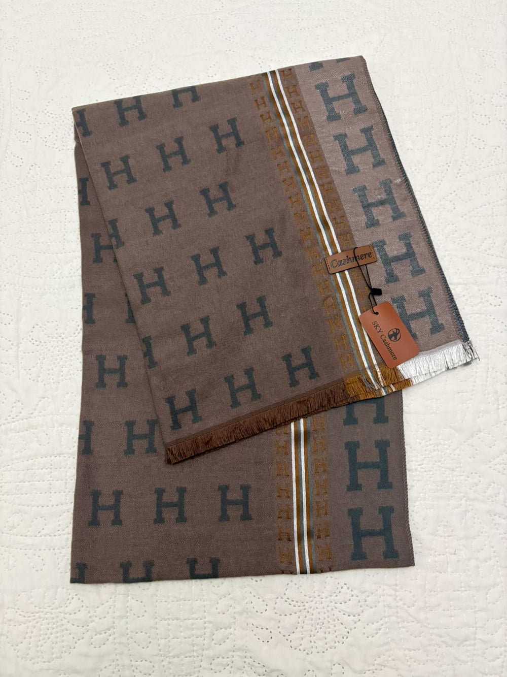 A folded Hermès scarf in taupe brown, featuring a repeating dark blue 'H' monogram pattern, gold vertical stripes, and a fringed edge with a branded hangtag.