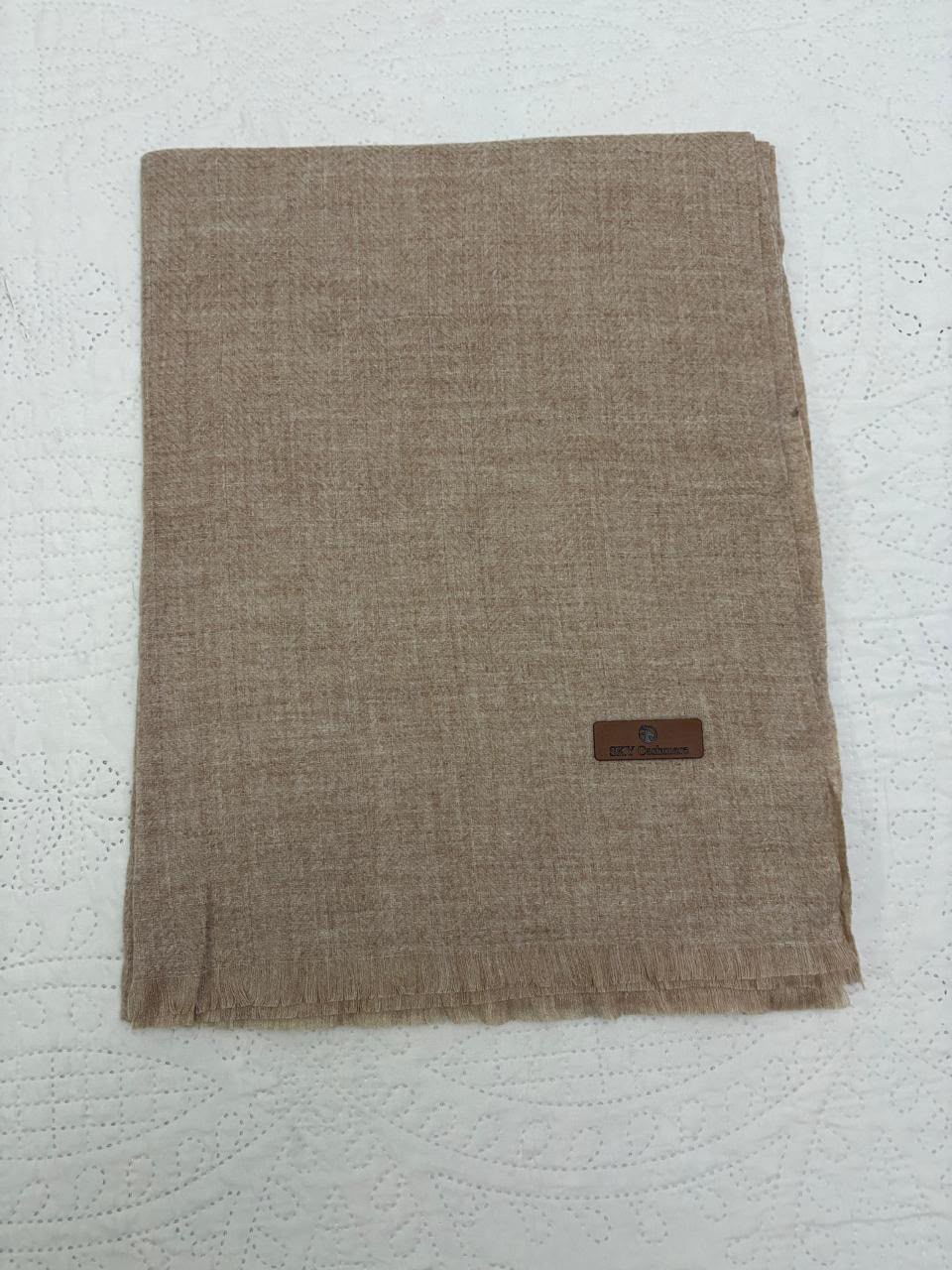 A folded beige woven scarf with a small brown leather brand tag, displayed on a white textured fabric background.