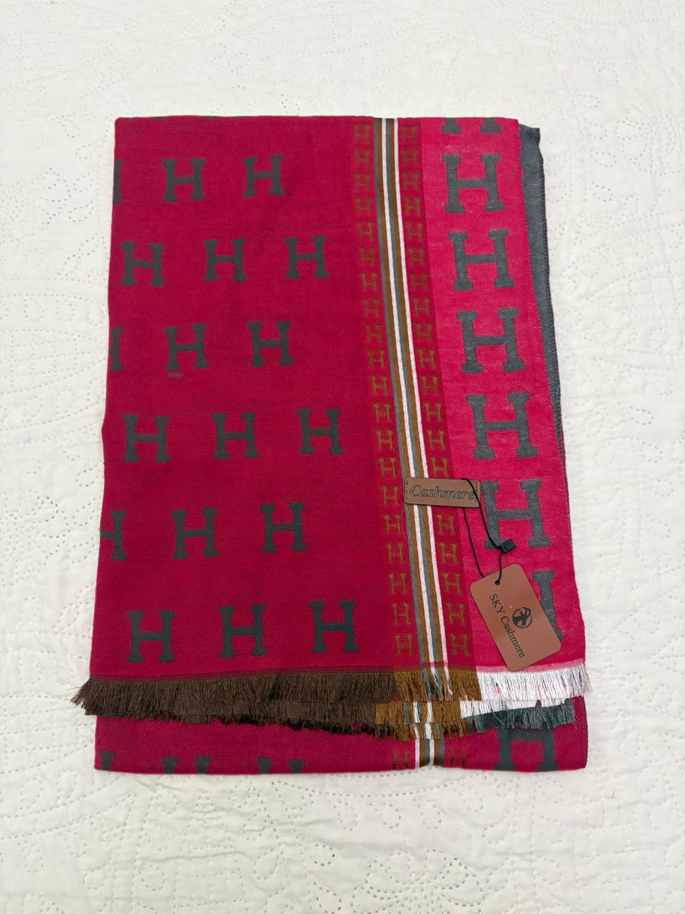 A folded pink scarf with a repeating dark green 'H' pattern, a striped border, and a brown fringed edge, shown on a white textured background with a hangtag attached.
